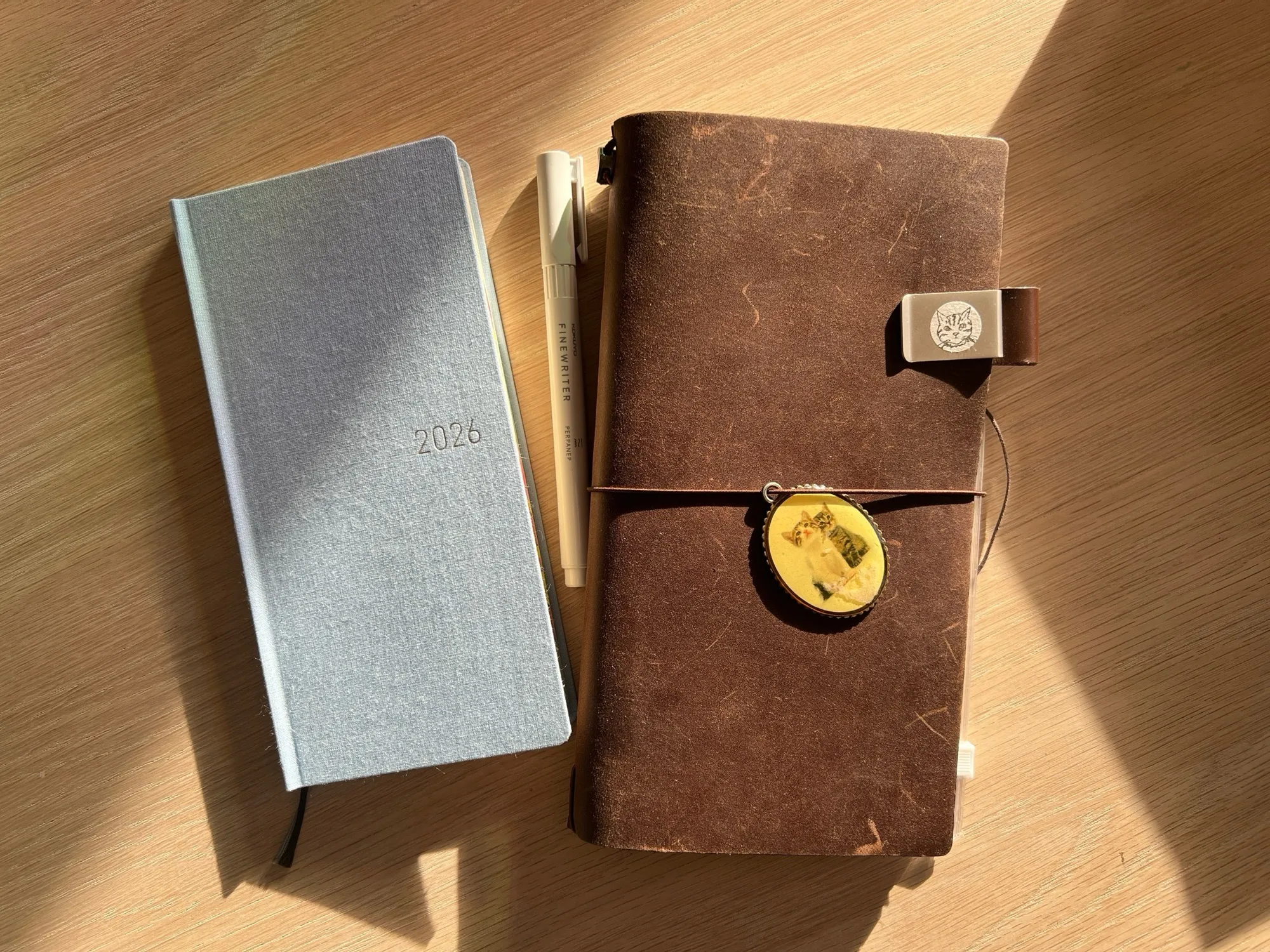 A grey 2026 Hobonichi Weeks and a pen next to a brown leather Traveler’s Notebook, on a sunny wooden desk